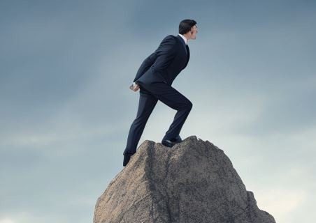 A man in a suit stands triumphantly on a rock surrounded by papers.