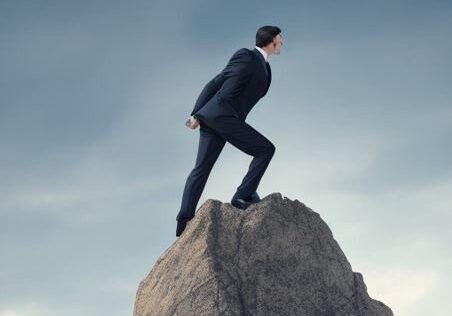 A man in a suit stands triumphantly on a rock surrounded by papers.